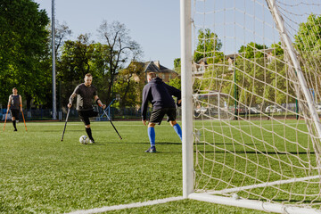 A man on crutches is dribbling a ball towards a goal in front of which a man is standing