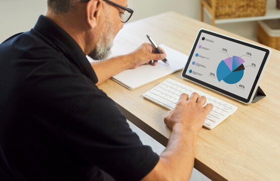 Man using tablet to analyze pie chart for financial accounting, business planning. Busy senior office worker monitoring metrics on digital screen, writing notes at workplace for data research - Powered by Adobe