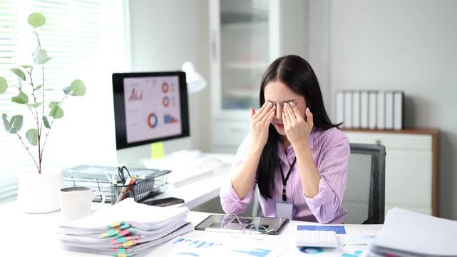 Overworked asian businesswoman feeling tired and suffering from eye strain, taking off her glasses and massaging her eyes while sitting at a messy desk with piles of documents