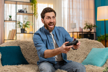 Young man sitting at home on sofa, gripping gamepad joystick, intensely playing video game with focus and excitement. Caucasian guy reacting emotionally to gameplay, immersed in virtual world, indoors