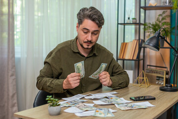 Middle-aged man at home reviews bills counts cash piles feeling anxious about high costs rising. Young adult guy at table sorts money with papers calculating debt worrying about future payment stress