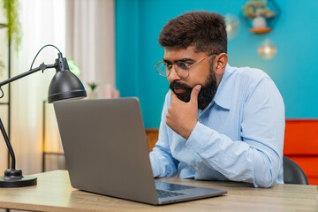 Indian man at home types on laptop stops rubs chin thinking over vital idea creative future plan. Arabian guy at table gazes screen focused eyes show deep concentration solving problem quickly today.