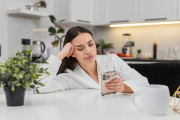 Young woman at home scrolls news on smartphone bored unhappy low energy emotional exhaustion burnout mood. Caucasian girl on sofa sits at table reads feed time slow thumb move tired eyes needs break.
