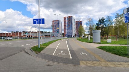 A sidewalk and a bike path with markings are laid out between the roadway and the park. Nearby there are lawns with grass, traffic lights, buildings and parked cars. Cloudy spring weather