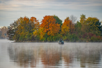 Fisherman in small boat on foggy Daugava River in Riga with autumn forest reflections creating calm scenic landscape of nature, travel and seasonal balance in early morning light