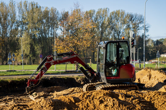 Compact red mini excavator performing ground excavation at construction site with soil mounds, hydraulic arm extended for trenching and groundwork preparation