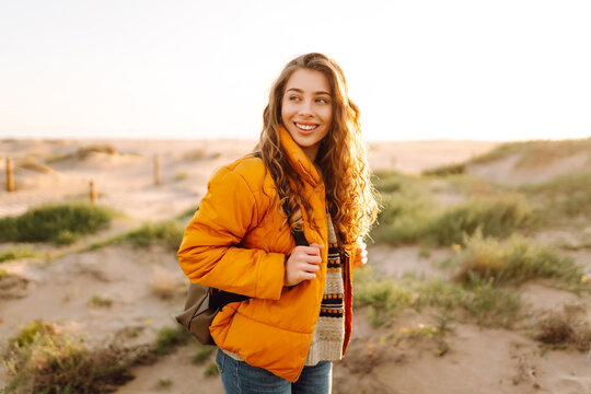 A young woman in a bright jacket walks along a sandy beach at sunset. A beautiful backpacker enjoys nature and an outdoor adventure. Concept of travel, nature, and relaxation.