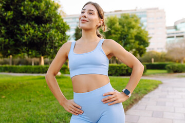 Fototapeta premium A young woman in athletic clothing enjoys a sunny day in a park. An athletic woman feels elated after a run and stretches outdoors. Concept of sport, enjoyment, and relaxation.