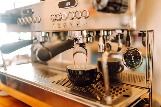 Experienced barista prepares freshly brewed coffee using professional coffee machine. In cozy coffee shop, espresso is poured from espresso machine into black cup. Small business, beverage concept.