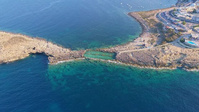 4K Drone aerial of a narrow coastal land bridge &mdash; thin isthmus connecting two larger landmasses, calm deep‑blue and turquoise water showing varying depths, road or pathway across the spit