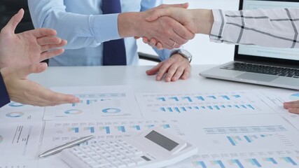 Business people shaking hands over financial documents and charts, confirming a successful agreement and partnership during a corporate meeting in the office