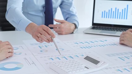 Business professional pointing at printed charts with a pen during a meeting, laptop showing bar graphs and calculator on table while team collaborates on financial analysis and planning - Powered by Adobe