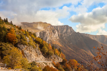 Spectacular mountain landscape with rocky cliffs, autumn forest, and clouds drifting across the peaks. Warm sunlight enhances the vibrant fall colors and natural beauty.