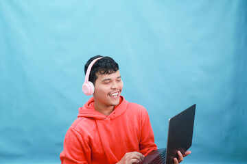 Young man engrossed in laptop with headphones against blue backdrop.