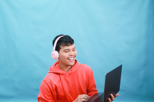 Young man engrossed in laptop with headphones against blue backdrop.