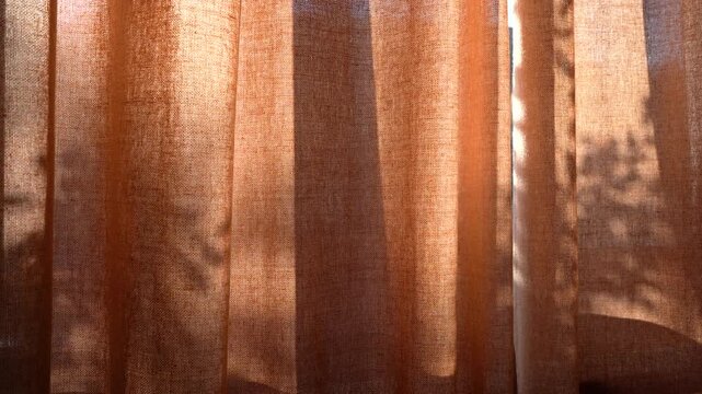 Warm morning sunlight filtering through textured, translucent orange curtains, casting soft shadows and creating a cozy, inviting atmosphere in a residential interior space