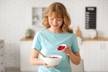 Beautiful young woman adding dehydrated raspberries into bowl with tasty granola at home