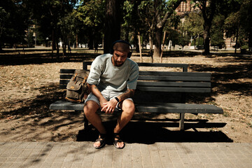Young man sitting on a bench in a sunny park with headphones and backpack. Concept of relaxation, freedom, and outdoor lifestyle