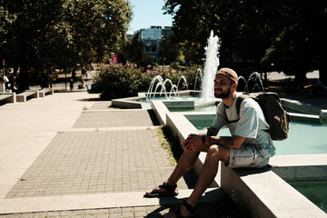 Young traveler with backpack sitting near a fountain enjoying sunshine and fresh air. Concept of freedom, leisure, and summer lifestyle