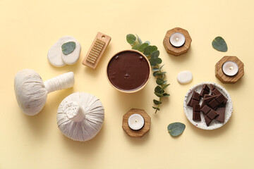 Spa composition with burning candles, herbal massage bag and bowl of melted chocolate on pale yellow background