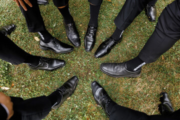 Groomsmen in polished black dress shoes and formal trousers stand in a circle on green grass, showcasing stylish footwear and unity before a wedding ceremony celebration