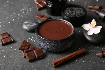 Spa composition with bowl of melted chocolate, pieces of chocolate bar and cinnamon stick on black background, closeup