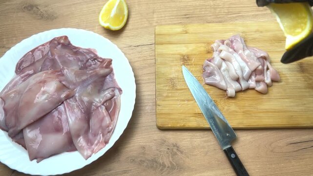 Close-up view from above on a wooden cutting board there are peeled squids cut into rings, the hand of a chef in a black glove pours them with juice from a lemon slice, next to it is a plate with