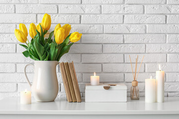 Burning candles, books and tulip flowers in vase on table near white brick wall in room, closeup