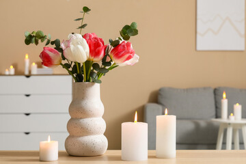 Beautiful flowers in vase and burning candles on table in interior of living room, closeup