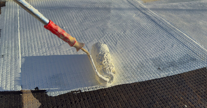 Worker applying white polyurethane waterproof coating on flat roof surface with paint roller.