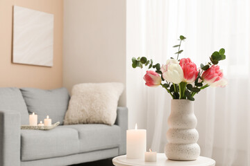 Beautiful flowers in vase and burning candles on table in interior of living room, closeup