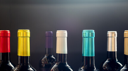 Striking, low-key, close-up photograph of the necks of multiple dark wine bottles lined up, distinguished by their vibrant and diverse colored foil capsules against a dark, dramatic background