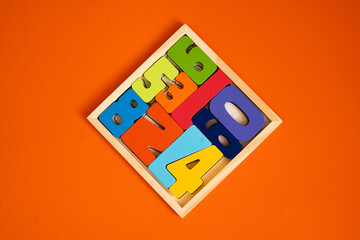 A wooden puzzle tray filled with colorful wooden numbers sits on a bright orange background. The pieces, in blue, green, yellow, red, and orange, fit neatly inside a square wooden frame.