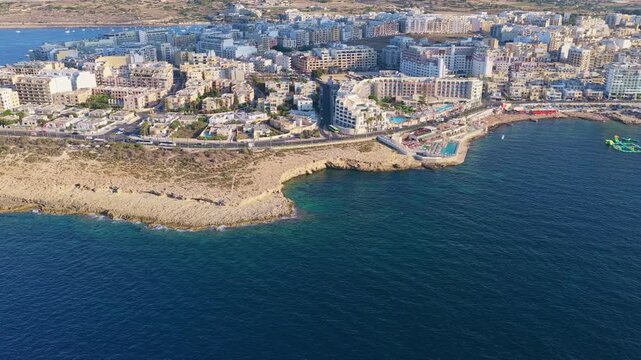 4K Drone aerial of dense coastal cityscape &mdash; rocky shoreline with small piers, mid‑ to high‑rise buildings, active roads and deep‑blue sea under clear daytime skies for urban and coastal B‑roll