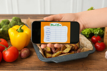 Young woman using meal plan app on mobile phone and vegetables on table in kitchen, closeup