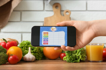 Young woman using meal plan app on mobile phone on wooden table in kitchen, closeup