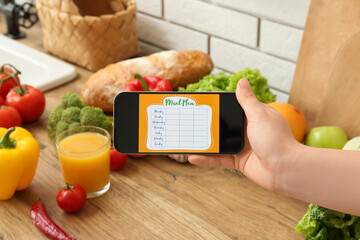 Young woman using meal plan app on mobile phone on wooden table in kitchen, closeup