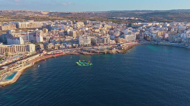 4K Drone aerial of a coastal city with floating water-park &mdash; dense urban shoreline, roads and recreational areas bordering clear sea, bright green inflatable play structures anchored near shore