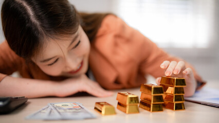 Asian adult woman happily planning wealth investment with gold bars and cash on desk at home office achieving financial success growth prosperity