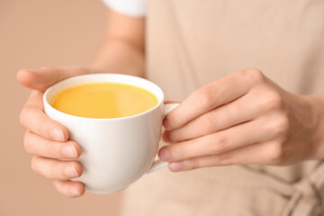 Young woman holding cup of tasty turmeric drink on beige background, closeup
