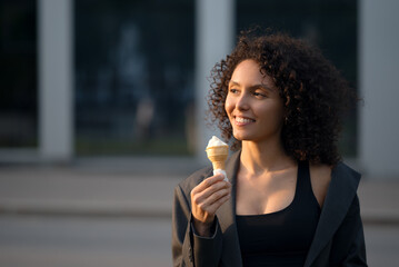 Smiling young woman with curly hair in a gray blazer holding ice cream outdoors at sunset with copy space, representing modern summer lifestyle, elegance, and positive emotion.