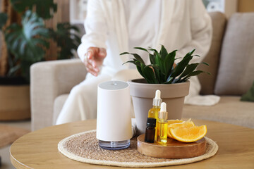 Air humidifier with essential oils, orange and plant on table against woman sitting at home, closeup