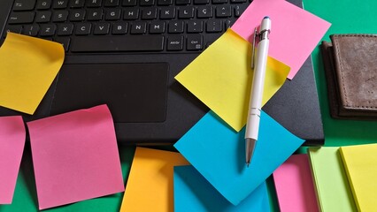 Desk essentials with laptop, colorful post-its, pen and leather wallet on green background