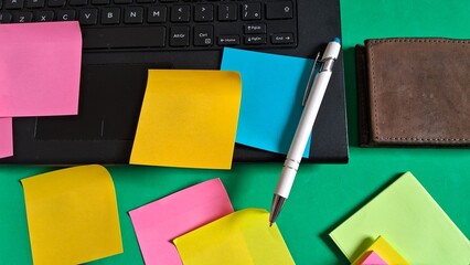 Desk essentials with laptop, colorful post-its, pen and leather wallet on green background