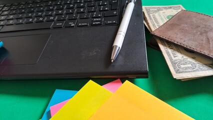 Desk essentials with laptop, colorful post-its, pen and leather wallet with dollars inside on green...