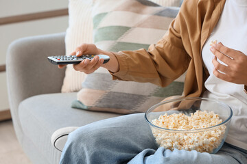 Young woman with remote controller and popcorn watching TV on sofa at home, closeup