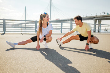 Two people stretching in workout clothes on a waterfront area with a bridge in the background