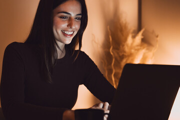Head shot pleasant happy young woman freelancer working on computer at night. Attractive businesswoman studying online, using laptop software, web surfing information or shopping in internet store.