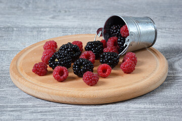 The natural fresh raspberry and blackberry on the wooden plate with a decorative metallic bucket.