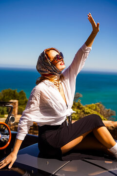 Stylish woman in sunglasses and silk scarf posing on a vintage convertible car, raising her hand with a smile, enjoying a sunny day by the sea.